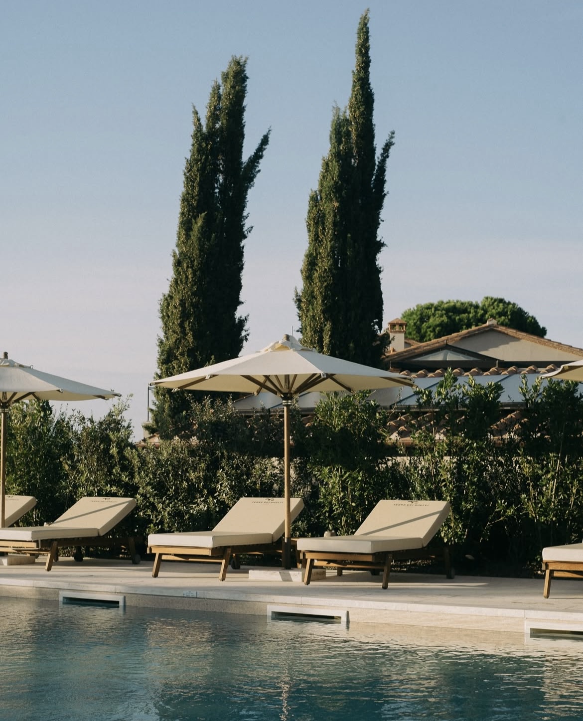 Post Instagram della piscina dell'agriturismo toscano Bolgheri Terre dei Ghelfi: sullo sfondo due cipressi, avanzando bordo piscina con lettini e ombrelloni. In primo piano porzione di piscina.